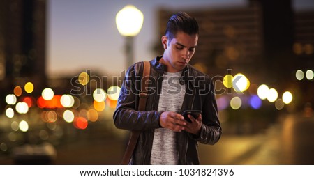 Millennial Hispanic man standing on city bridge at night looking at smartphone Royalty-Free Stock Photo #1034824396