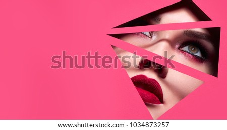 the face of a young beautiful girl with a bright make-up and with plump red lips peeks into a hole in pink paper