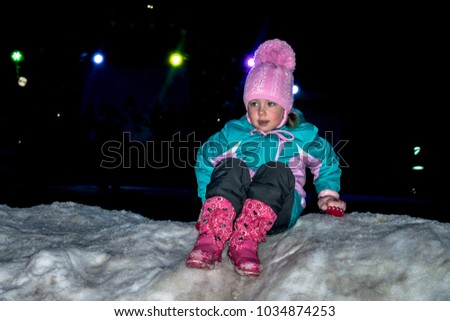 A little girl climbs with a snow slide at night. A child is playing on the ice in the dark. The baby is sitting on the snow. Winter children's entertainment
