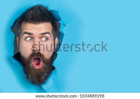 View of male face through hole in blue paper. Surprised bearded man making hole in paper. Copy space for advertising, to insert text or slogan. Discount, sale, season sales. Royalty-Free Stock Photo #1034889298