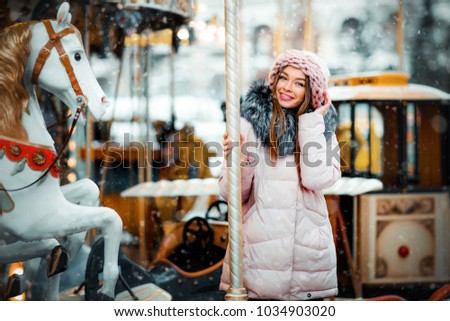 Portrait of a beautiful young model in pink knitted hat standing near carousel on a christmas background with bokeh lights. Magic warm new year photo.Cozy background. Happy woman.