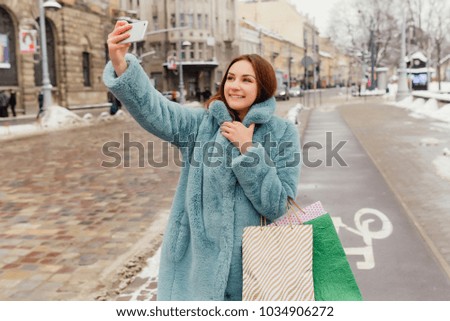 Selfie photo of smiling attractive girl with paper bags after shopping. Laughing pretty woman wearing fur coat in winter time make selfie portrait.