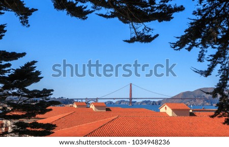 A view of the Golden Gate Bridge from the heights above Fort Mason in San Francisco. Trees frame the picture of red tile roofs and a view of the bay. A blue sky is in the background.
