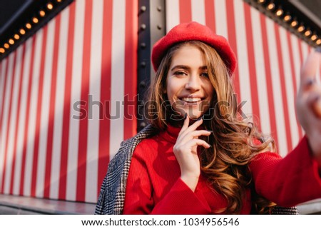 Inspired pale girl with shiny curly hair taking picture of herself on striped background. Cute female model in red knitted clothes having fun and making selfie.