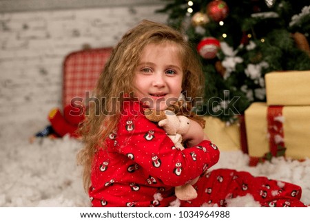 Curly-haired girl in red pajamas happily hugs her favorite stuffed toy, Christmas background