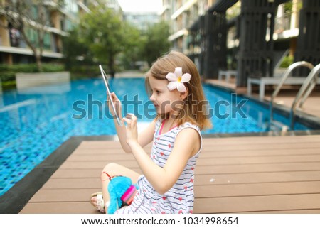 kid girl uses tablet sitting close to blue swimming pool wearing flower and hold toy. concept of new technologies travel, recreation or holiday family time on sunny day
