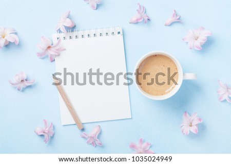 Notebook, cup of coffee and pink flowers on blue pastel background top view. Fashion woman working desk. Spring to do list. Flat lay style.
