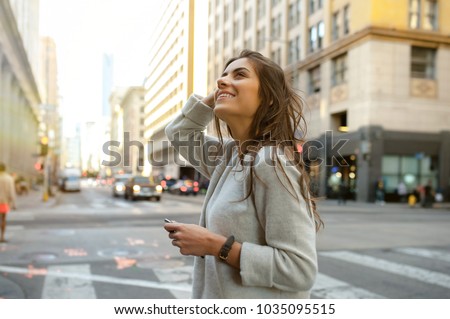 Beautiful young woman on the boulevard in urban scenery, downtown, at sunset, holding smartphone and looking side.