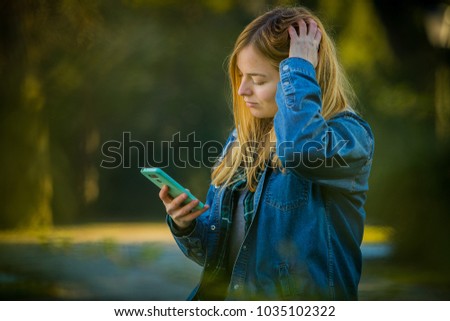 Cute blonde girl with long hair, septum piercing and jeans shirt using her telephone as a mirror in a park with strong sunny backlight. Looking towards the phone and holding her hair with one hand.
