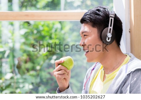 Side view close-up portrait of a young man eating a fresh green apple while listening to stereo headphones indoors