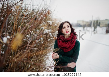 Brunette girl in green sweater and red scarf outdoor against bushes on evening winter day.