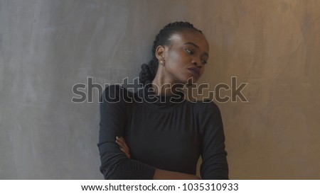 Depressed beautiful african woman with pigtails, purple lipstick and arms crossed is leaning on the gray wall and looking aside.