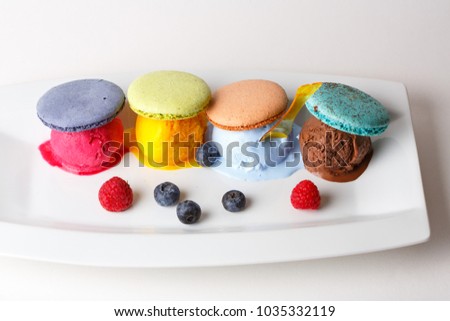 Rainbow colored ice cream and macaroons set on white plate with berries Royalty-Free Stock Photo #1035332119