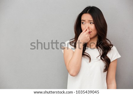Portrait of frustrated woman 20s pinching nose with disgust on his face due to bad smell isolated over gray background Royalty-Free Stock Photo #1035439045