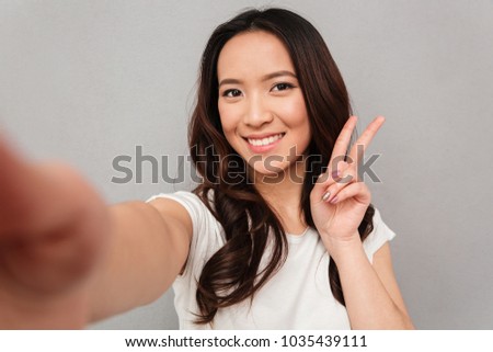 Beautiful woman with asian appearance taking selfie and showing victory sign with perfect smile isolated over gray background
