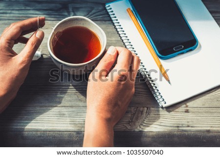 Woman hands on wooden table with cup of tea,paper notepad,phone. Dark vintage toned image.