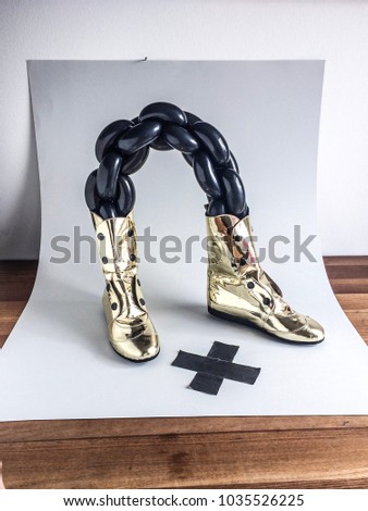 high-top shoes with gold mirror finish connected with matte black balloon chain next to black duck tape cross mark, isolated on white background on wooden table, conceptual photography style
