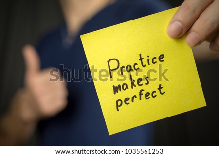 Man holds yellow sticker inscription Practice makes perfect. Thumb up Blue T-shirt. close-up, selective focus