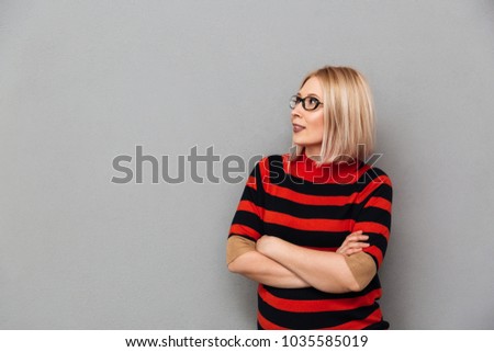 Smiling middle-aged blonde woman in sweater and eyeglasses looking away with crossed arms over grey background
