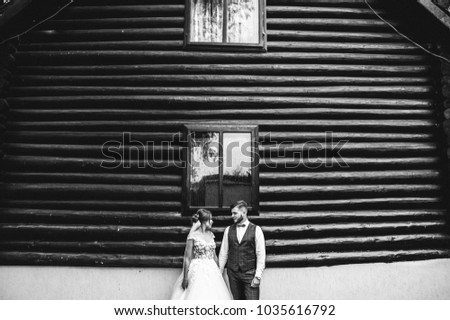 Beautiful wedding couple is standing near the wooden house. The bride with tulle veil and elegant hairdo is hugging her bearded groom in bow tie. Rustic outdoors stylish love story. Black and white.