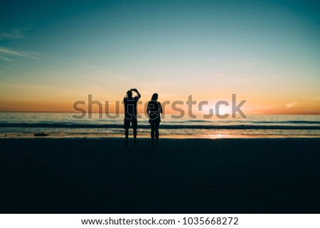 Silhouettes of male and female enjoying  scenic sunset, young couple in love enjoying beauty of evening tropical island during summer vacations honeymoon while taking picture on mobile phone