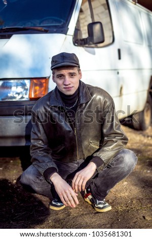 A young guy of criminal appearance in a black leather jacket stands near an old white van.