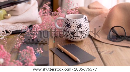 Aramatic coffee in a mug with a cat. Planning the day in notepad. Decor of pink flowers and a hat with glasses. A cozy house. Free space for text. Card.