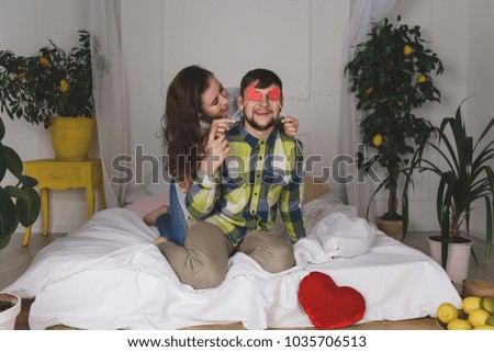 I love you. Happy young woman is covering female eyes with surprise and giving her a heart card. They are sitting on bed and smiling