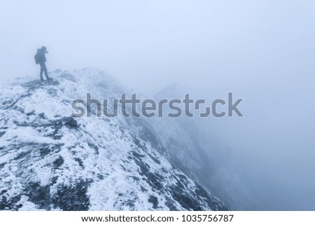 A tourist photographer at the edge of a snow-covered rock in a fog takes pictures