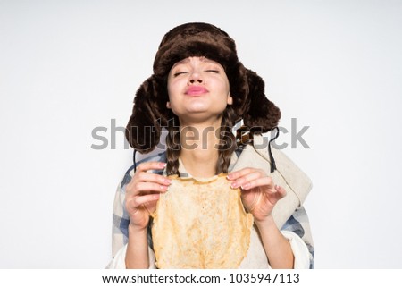 a funny young girl from Russia, wearing a warm fur hat, celebrating a carnival, holding a hot pancake