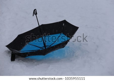 Outdoor view of a blue and black open umbrella located on the snowy ground. Isolated element on a white surface. Abstract minimalist image of a colorful object. Single accessory in a funny situation. 