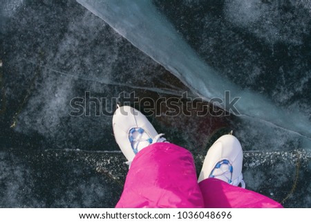 Legs of the girl in skates and pink winter warm pants on the beautiful ice of Lake Baikal with cracks and snow in winter on a sunny day.