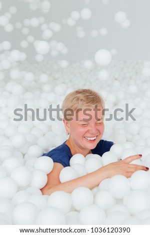 Portrait of beautiful senior woman with white short hair surrounded by white plastic balls in the dry pool. Copy splace. Adult childhood at Playground with pit-ball. Rain from falling balls.
