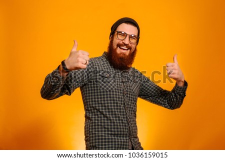 Portrait of cheerful happy bearded man wearing eyeglasses and hat and showing thumbs up over orange background, studio shot. 