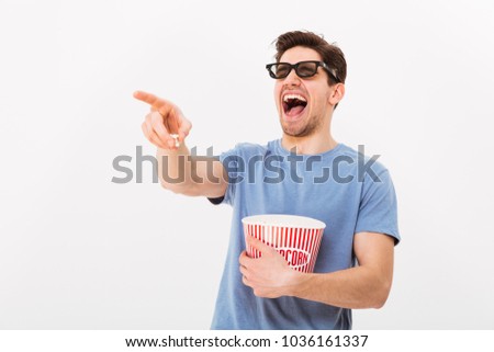Laughing man in t-shirt and 3d glasses holding bucket with popcorn while pointing and looking away over grey background