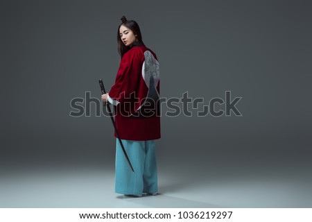 back view of samurai in kimono holding katana and looking at camera on grey   Royalty-Free Stock Photo #1036219297