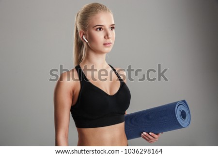 Portrait of a confident young sportsgirl listening to music with earphones while standing and holding fitness mat isolated over gray background