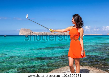 Pretty young female dressed in a coral clothes having fun making selfie with a selfiestick on the background of the azure sea