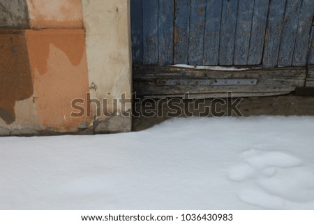Abstract outdoor view of a part of an ancient blue wooden door. Picture taken in the street during a snowy day. Orange and beige painted wall. Snow on the ground. Wet and aged textured surfaces.  