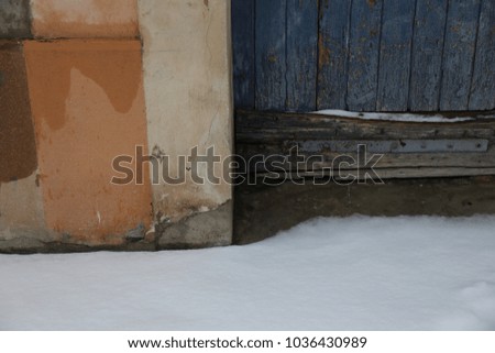 Abstract outdoor view of a part of an ancient blue wooden door. Picture taken in the street during a snowy day. Orange and beige painted wall. Snow on the ground. Wet and aged textured surfaces.  