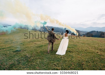 The bride and groom are running on the green hills with smokky sticks in their hands