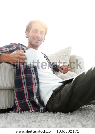 young man with laptop holding a cup sitting on the floor near th