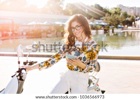 Lovely dark-haired lady wearing stylish white pantsuit and big headphones sitting on scooter and looking with shy smile. Outdoor portrait of trendy girl in elegant attire chilling near city fountain.