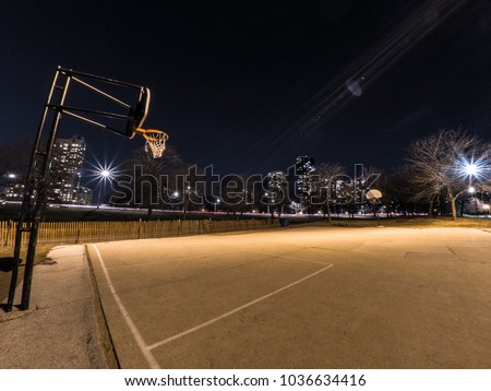Wide angle photograph of a concrete exterior basketball court with hoops at either end, street lights with residential highrise apartments in the distance in the dark night sky in Chicago.
