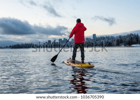 Adventurous male is paddle boarding near Stanley Park with Downtown City Skyline in the background. Taken in Vancouver, BC, Canada, during a vibrant winter sunrise.