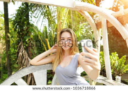 Young woman taking photo selfie on smart phone on sunny day, background of sunshine green palms in Thailand, Phuket. Concept of social networks, new technologies for better life, travel to tropical