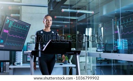 Beautiful Woman Athlete with Electrodes Connected to Her Body Walks on a Treadmill in a Sports Science Laboratory. In the Background High-Tech Laboratory with Monitors Showing EKG Readings. Royalty-Free Stock Photo #1036799902