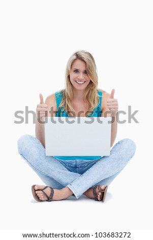 A young woman with her thumbs up is sitting on the ground with a laptop