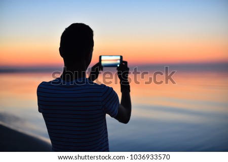 Silhouette of young guy taking a photo of a beautiful orange sunset over the Baltic sea with mobile phone camera. Summer morning before the sunrise. Dawn.