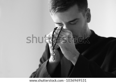Young priest with rosary beads praying on light background, toned in black and white